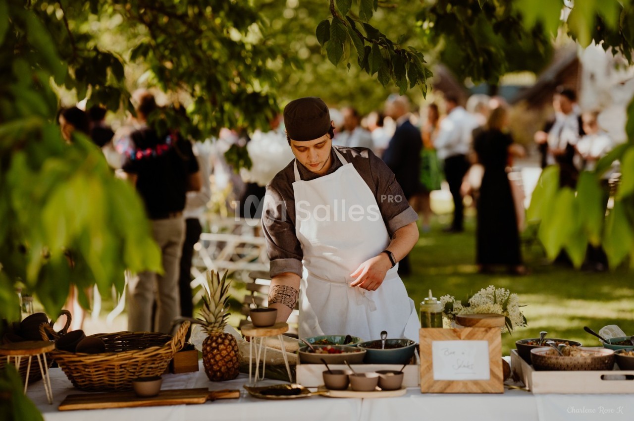 Un cuisinier prépare un buffet en plein air sous des arbres, avec des plats et des fruits disposés sur une table.