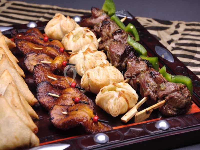 Assortiment de brochettes de viande grillée, beignets et samoussas sur un plateau en bois.