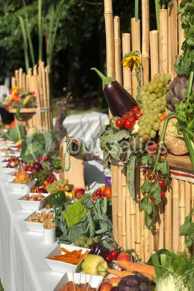 Buffet de légumes frais et fruits disposés sur une table décorée de bambous, en extérieur sous la lumière du soleil.