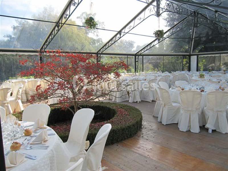 Salle de réception lumineuse sous une verrière, décorée avec des tables dressées et un petit arbre central entouré de…