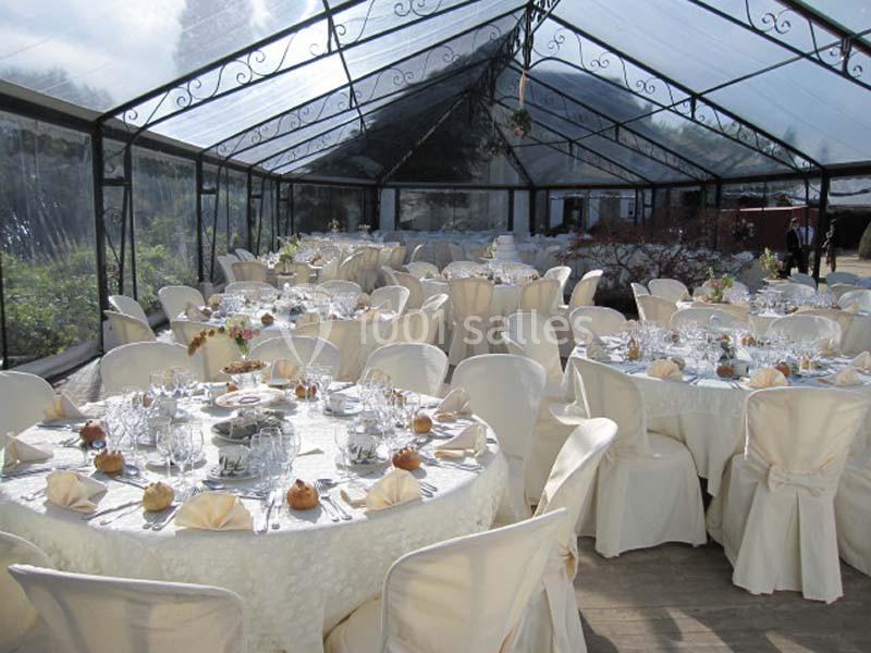 Salle de réception sous une verrière, tables rondes dressées avec nappes blanches, vaisselle et décorations élégantes.