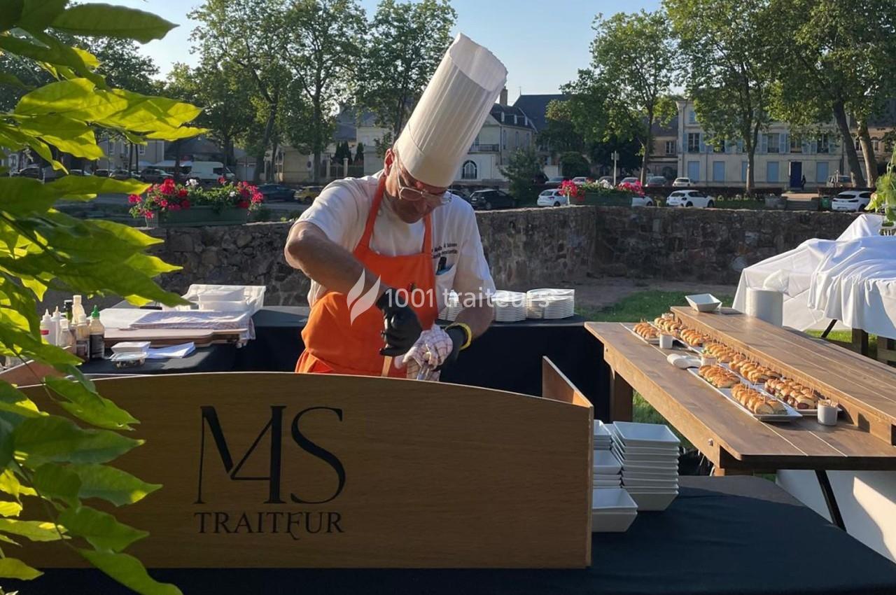 Un chef en tenue blanche et tablier orange prépare un plat derrière un comptoir en bois marqué ’M&S Traiteur’.