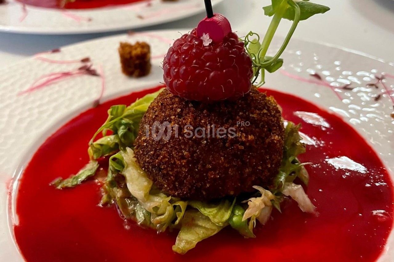 Boule croustillante sur lit de salade, garnie d'une framboise et décorée de sauce rouge sur assiette blanche.