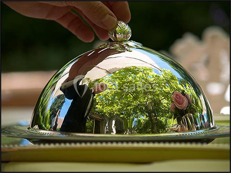 Traiteur Cloyes-les-Trois-Rivières (Eure-et-Loir) - Le Saint Jacques #2 Une main soulève un couvercle en métal réfléchissant, révélant un reflet d'arbres et de fleurs.