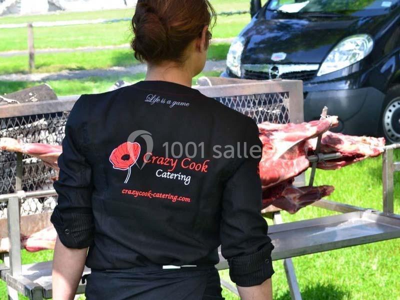 Une femme de dos en tenue professionnelle devant un agneau en cours de préparation sur une broche en extérieur.