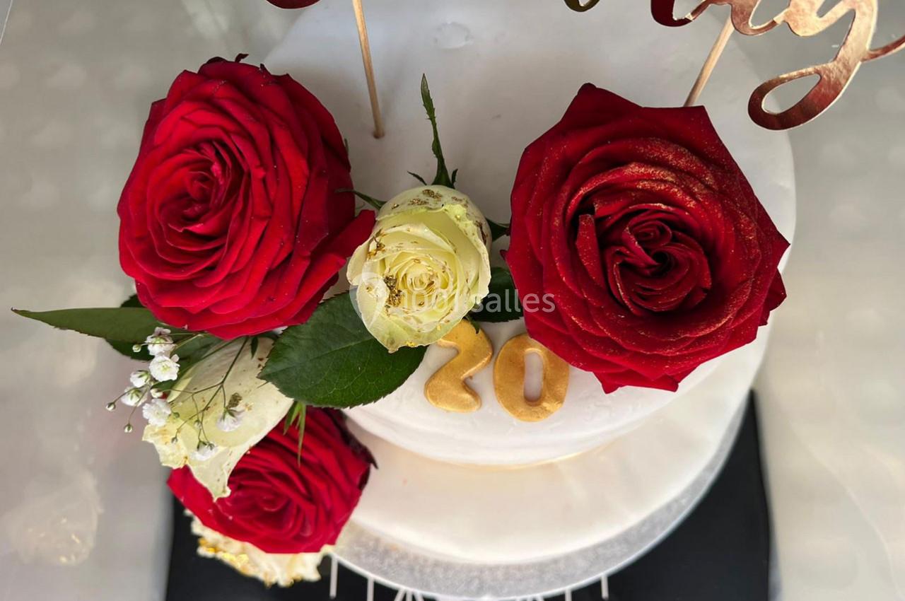 Gâteau blanc décoré de roses rouges et blanches, avec des chiffres dorés ’20’ et des éléments dorés en haut.