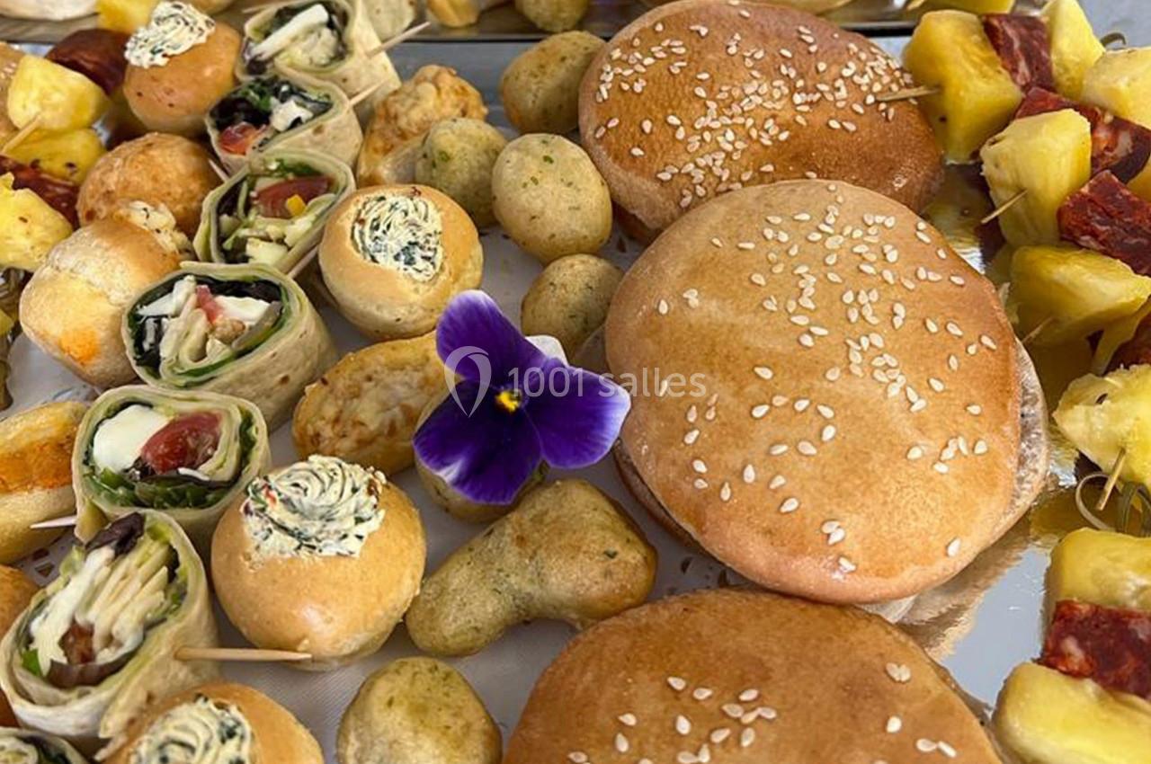 Assortiment de bouchées apéritives variées avec pains, roulés, brochettes de fruits et une fleur comestible violette.