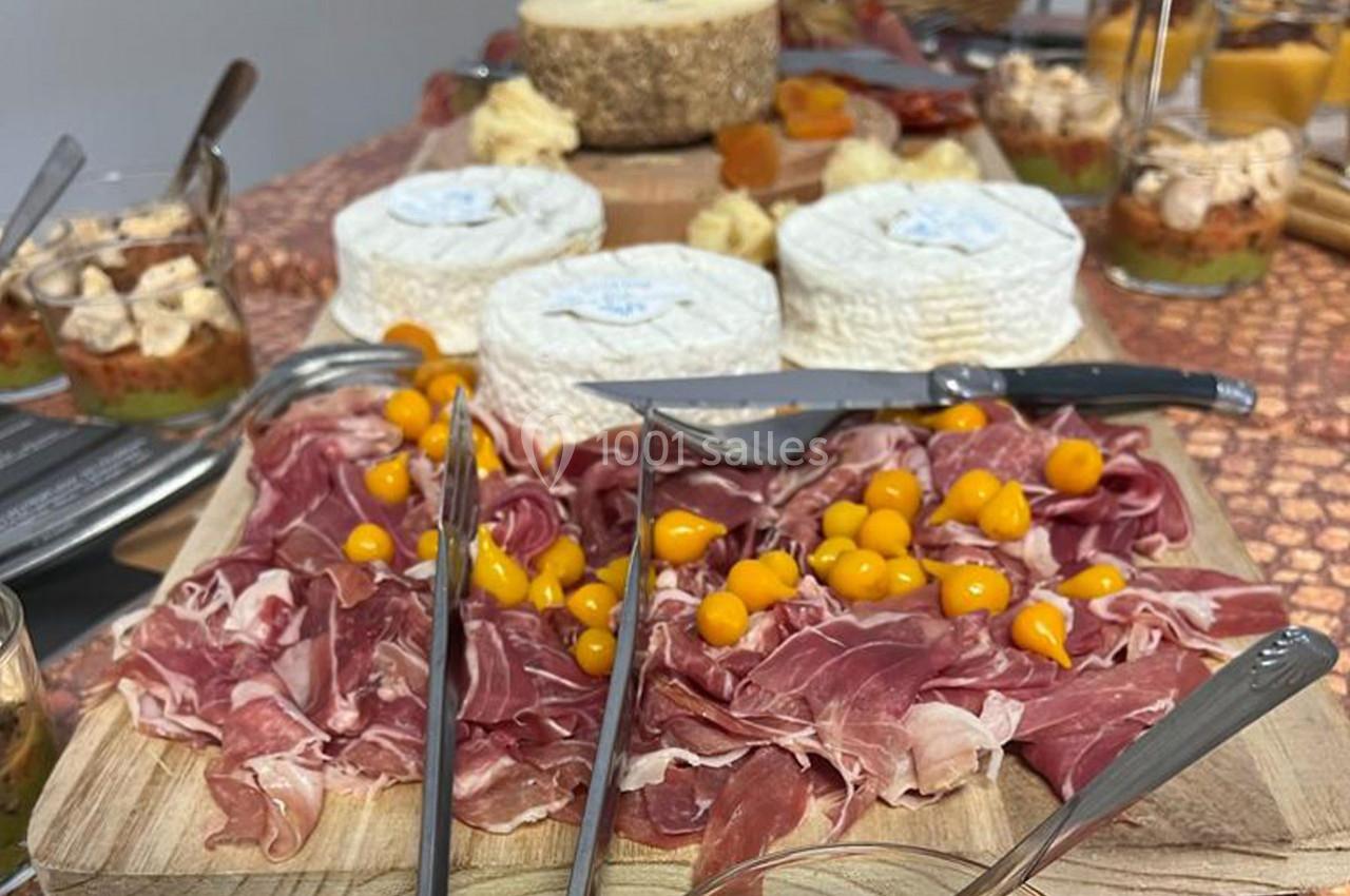 Plateau de charcuterie avec jambon cru et baies jaunes, accompagné de fromages et verrines en arrière-plan.