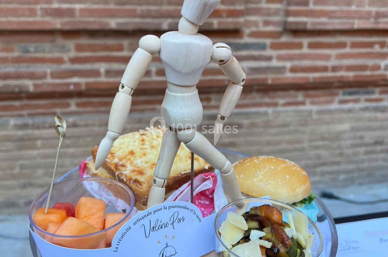 Un mannequin en bois posé devant des plats variés : salade, fruits, gratin et mini-burger, sur une table en extérieur.
