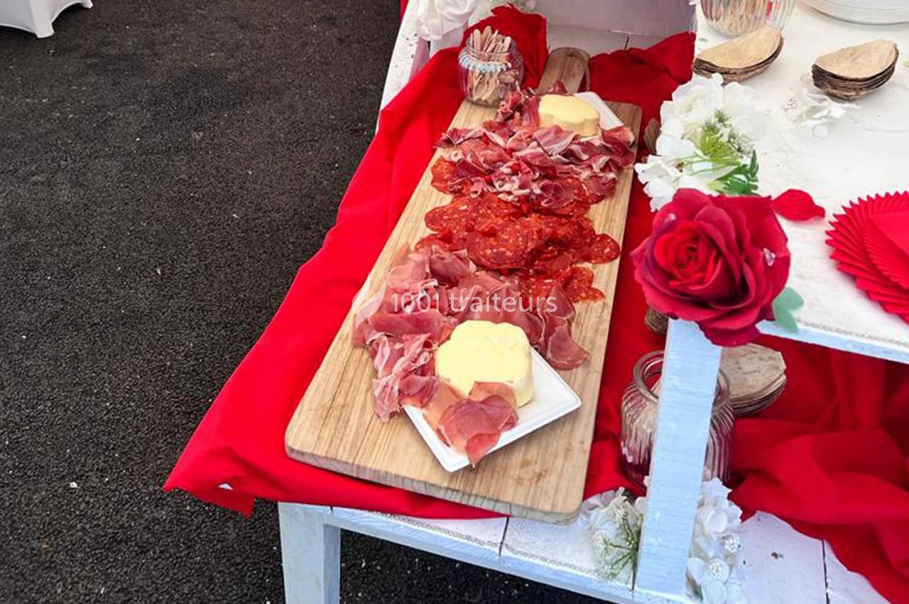 Plateau en bois garni de charcuterie, fromage et décoré de fleurs rouges sur une table blanche.