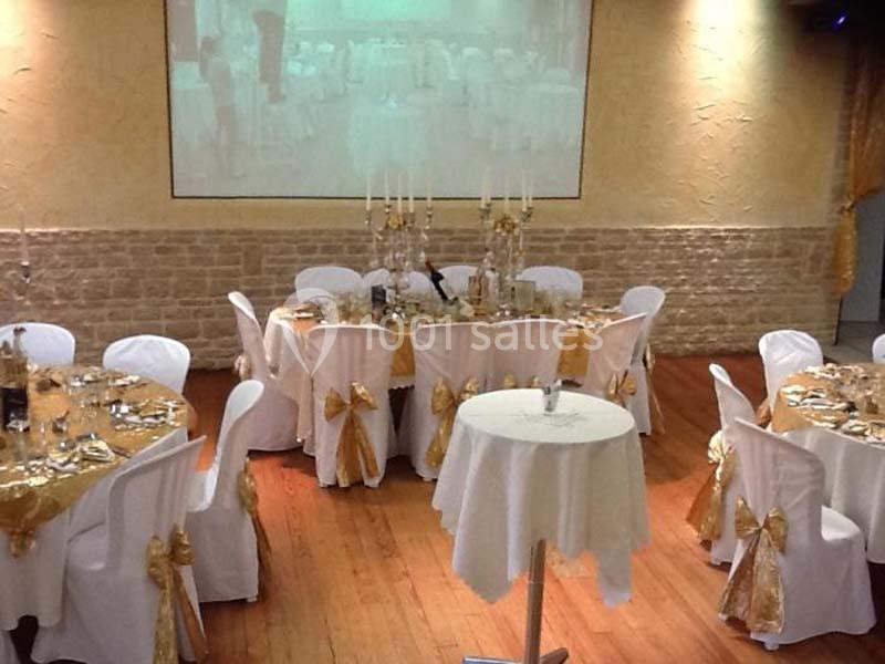 Salle de réception décorée avec des nappes blanches et dorées, tables rondes dressées et chandeliers centraux.