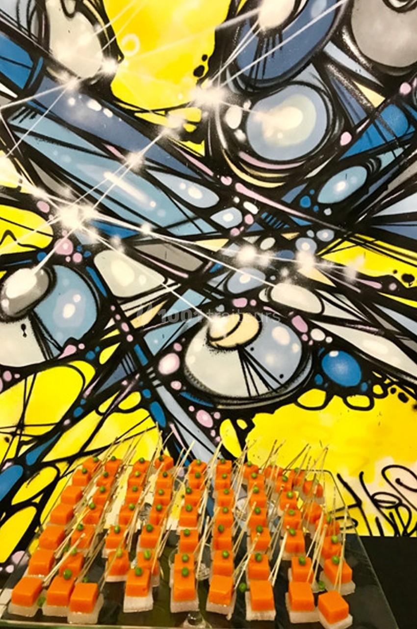 Bouchées apéritives colorées alignées sur un plateau devant une fresque murale abstraite jaune et bleue.