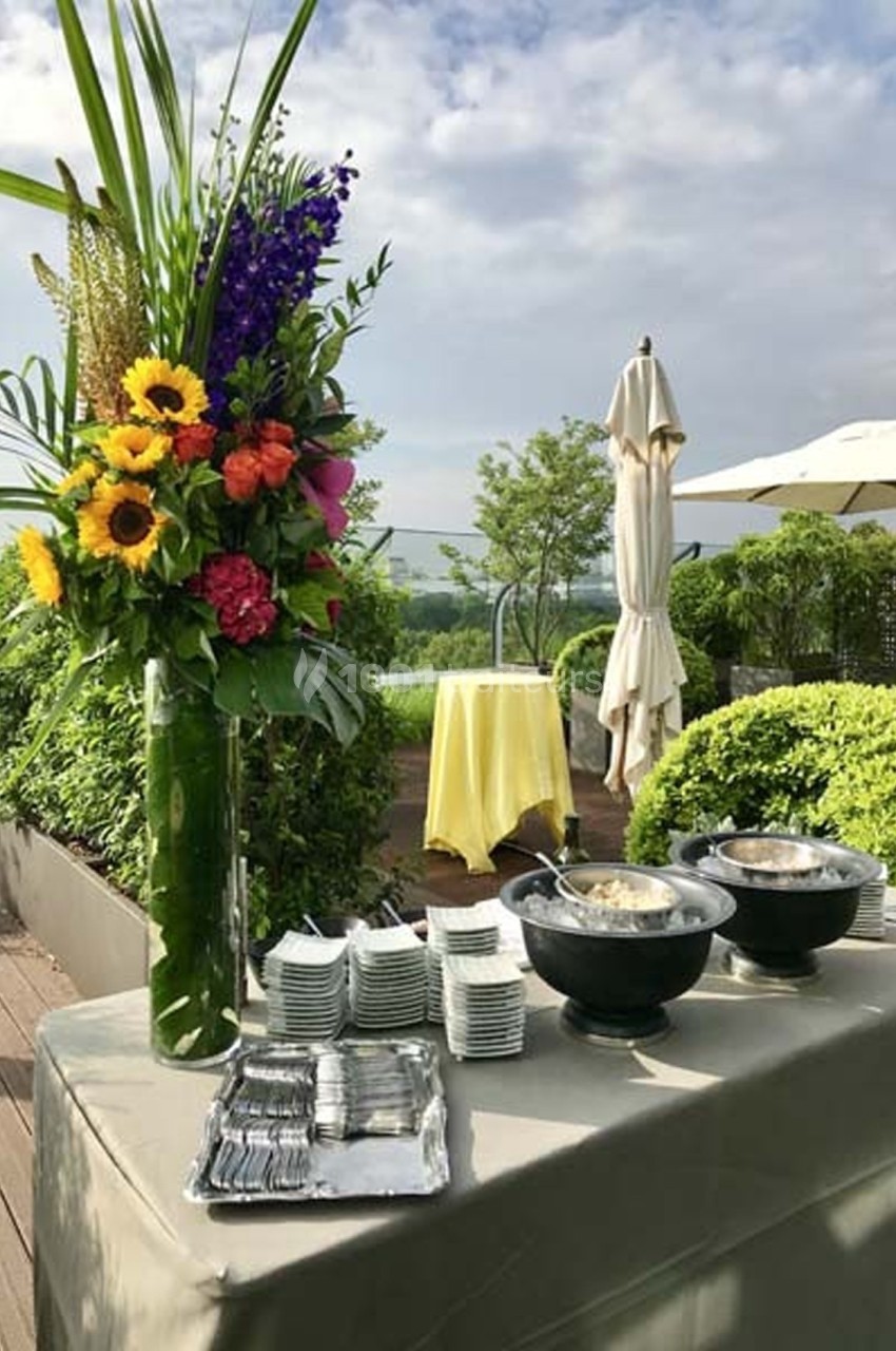 Table dressée en extérieur avec vaisselle, plats et un grand bouquet coloré, entourée de verdure et parasols.