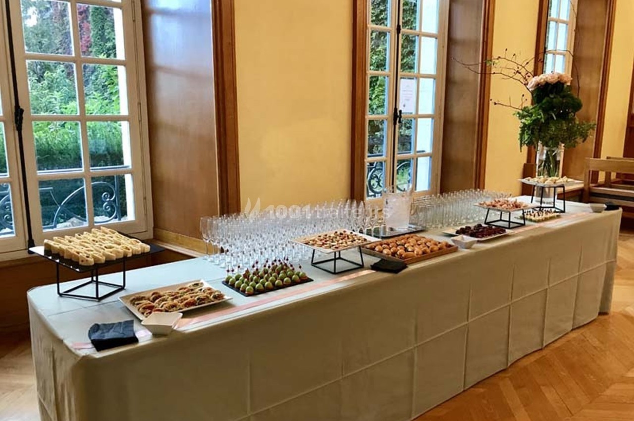 Table dressée avec des amuse-bouches variés, des verres vides et une décoration florale, dans une salle lumineuse.