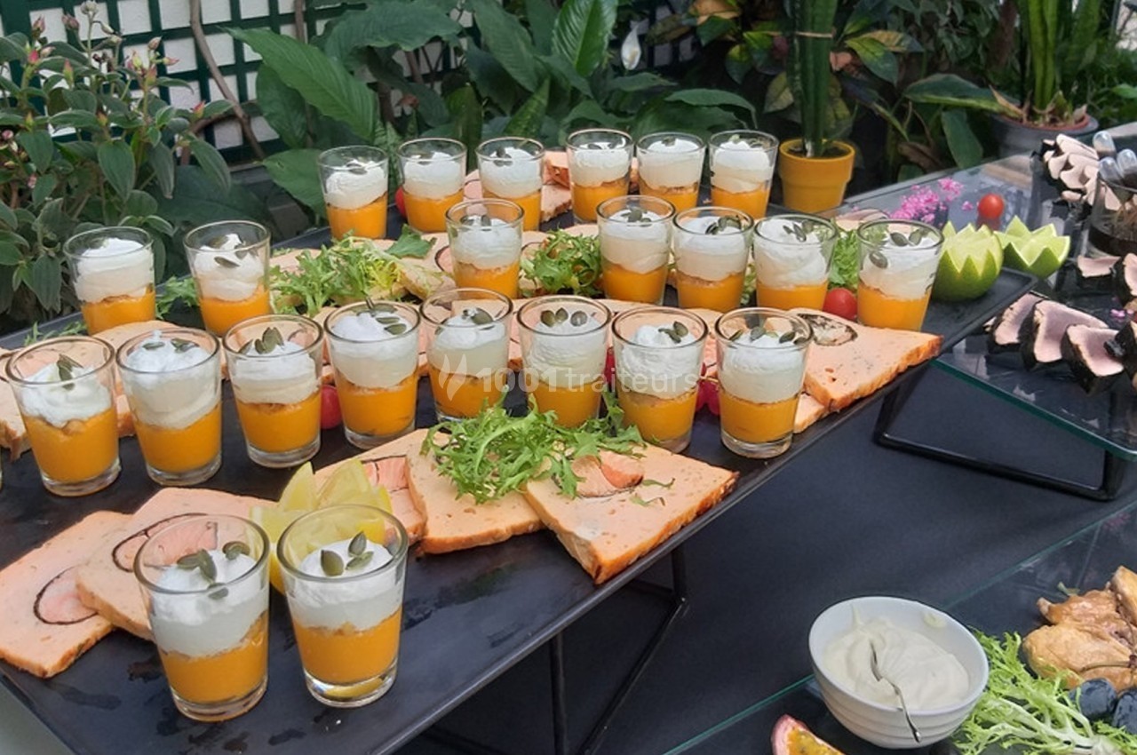 Buffet composé de verrines colorées, tranches de terrine et garnitures, disposé sur une table entourée de plantes.