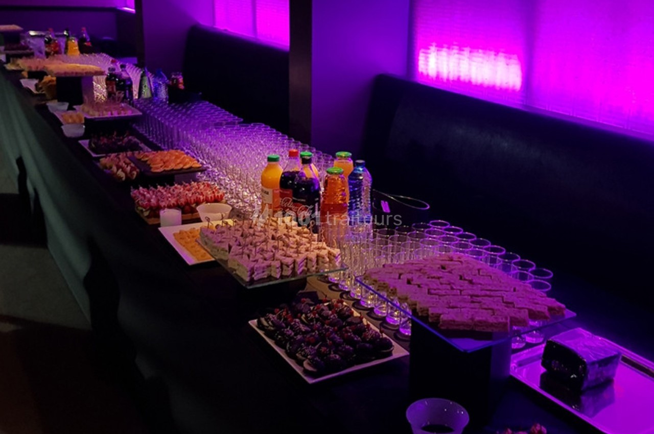 Buffet avec assortiments de boissons, verrines, canapés et desserts, disposé sur une table éclairée par une lumière violette.