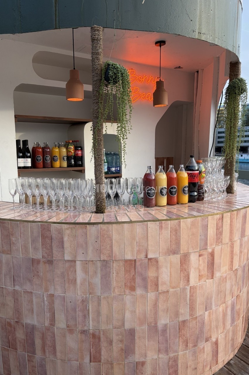 Bar extérieur arrondi avec carrelage rose, alignement de verres et bouteilles de jus devant un fond lumineux.