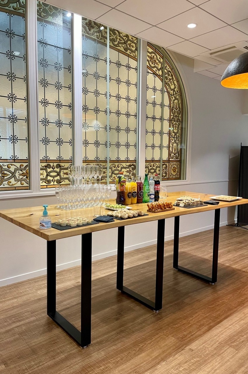 Table en bois avec verres, boissons, viennoiseries et désinfectant, située devant une verrière décorative.