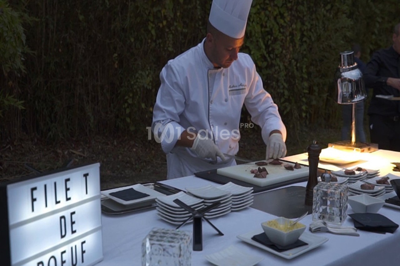 Un chef en tenue blanche découpe du filet de bœuf sur une table dressée en extérieur, éclairée par une lampe.