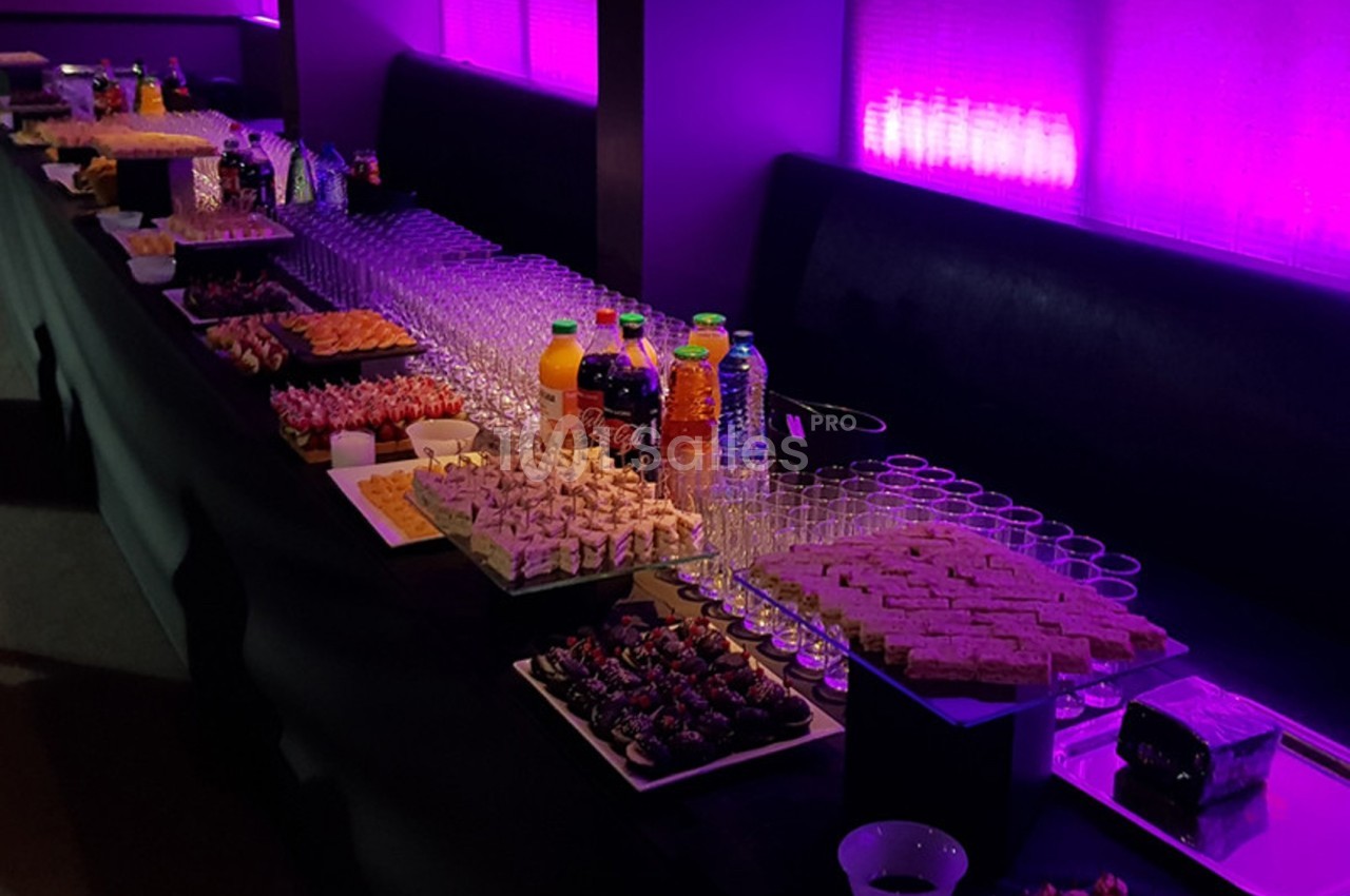 Buffet avec assortiments de boissons, verrines, canapés et desserts, disposé sur une table éclairée par une lumière violette.