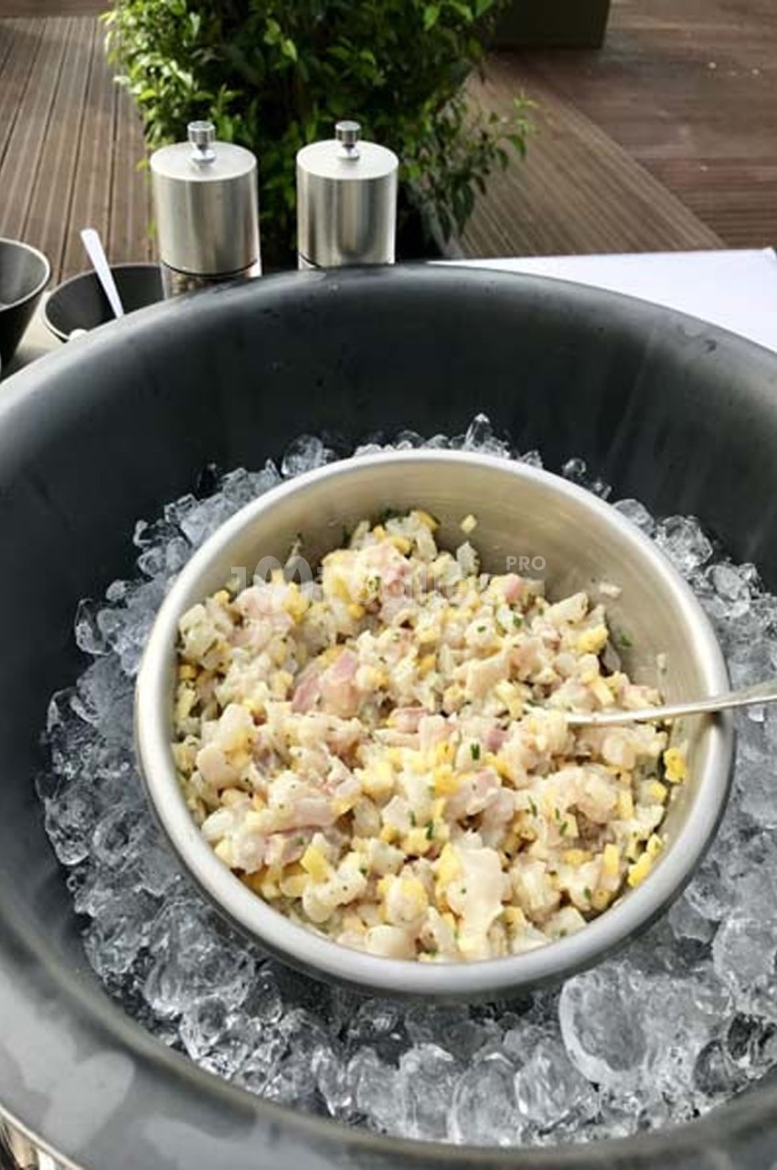 Bol en métal contenant un tartare de poisson cru, posé sur de la glace dans un récipient noir, avec des condiments en…