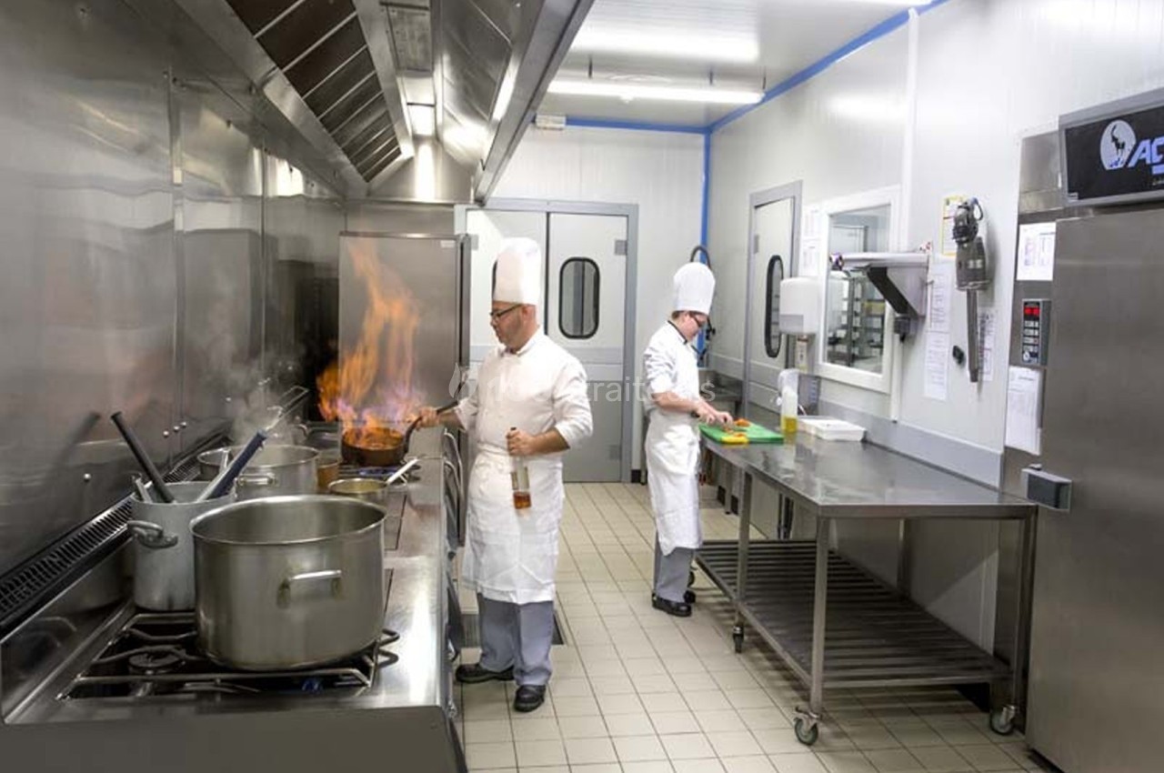 Deux chefs cuisiniers travaillent dans une cuisine professionnelle, l'un flambant un plat, l'autre préparant des ingrédients.