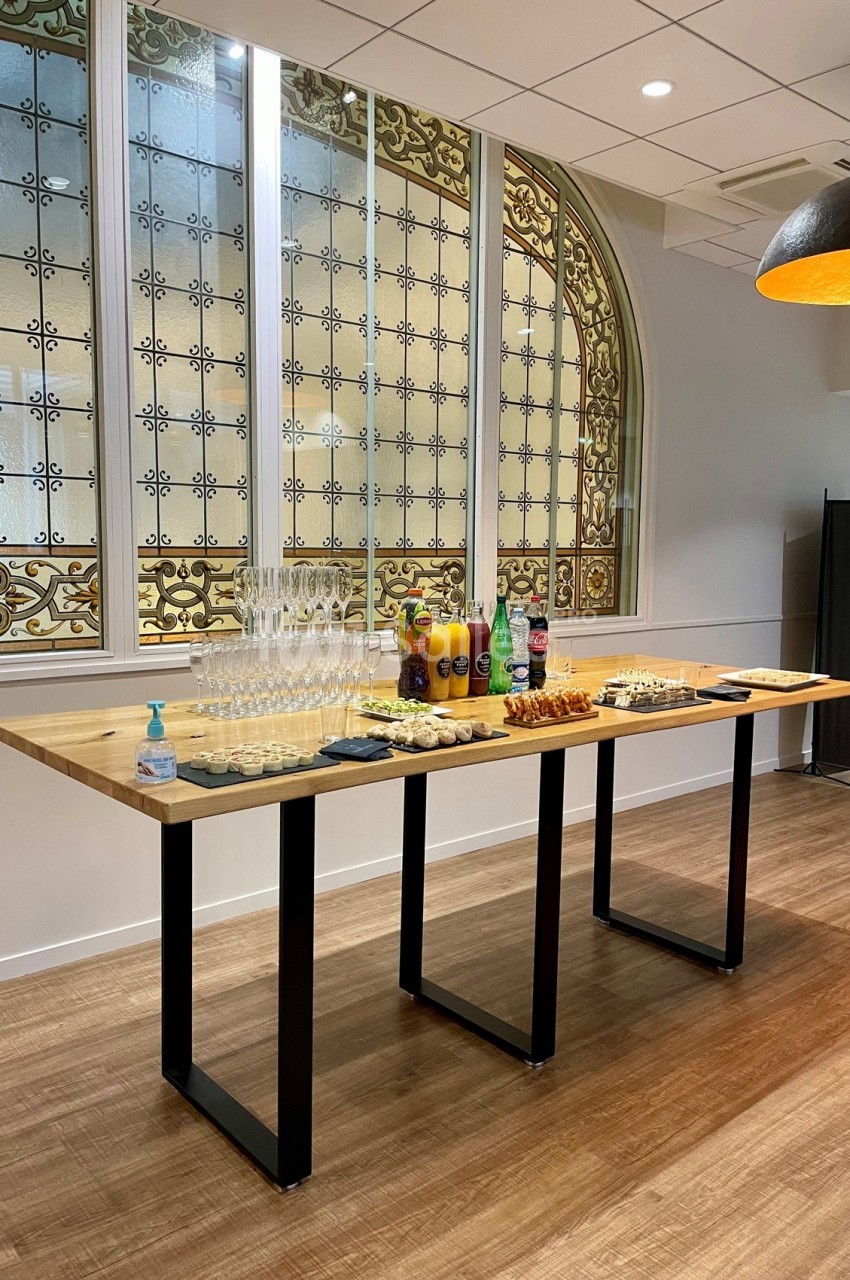 Table en bois avec verres, boissons, viennoiseries et désinfectant, située devant une verrière décorative.