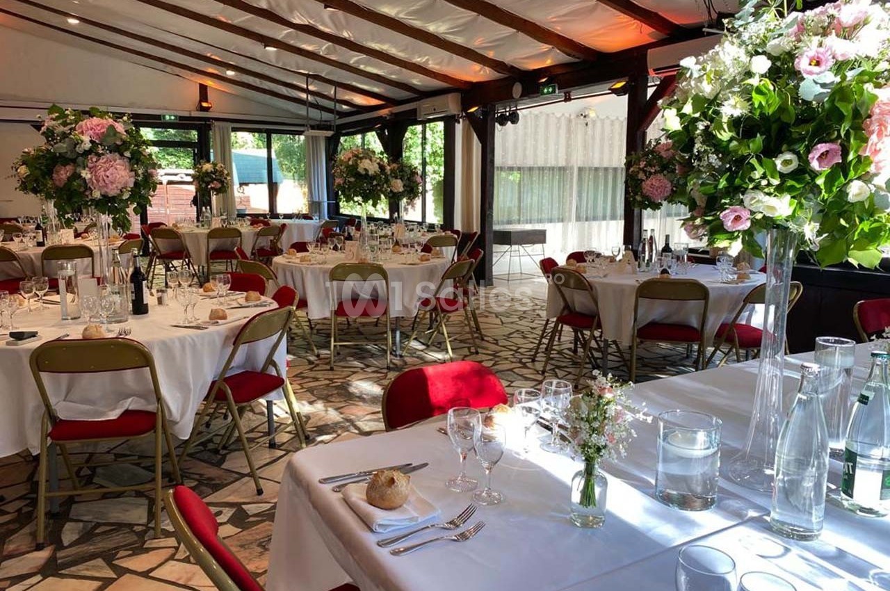 Salle de réception lumineuse avec tables dressées, chaises rouges et décorations florales élégantes.
