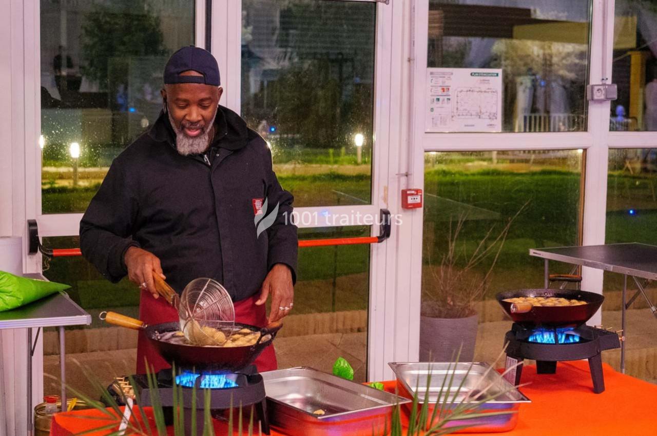 Un cuisinier souriant prépare des plats frits dans des poêles sur des réchauds, dans un espace lumineux et vitré.