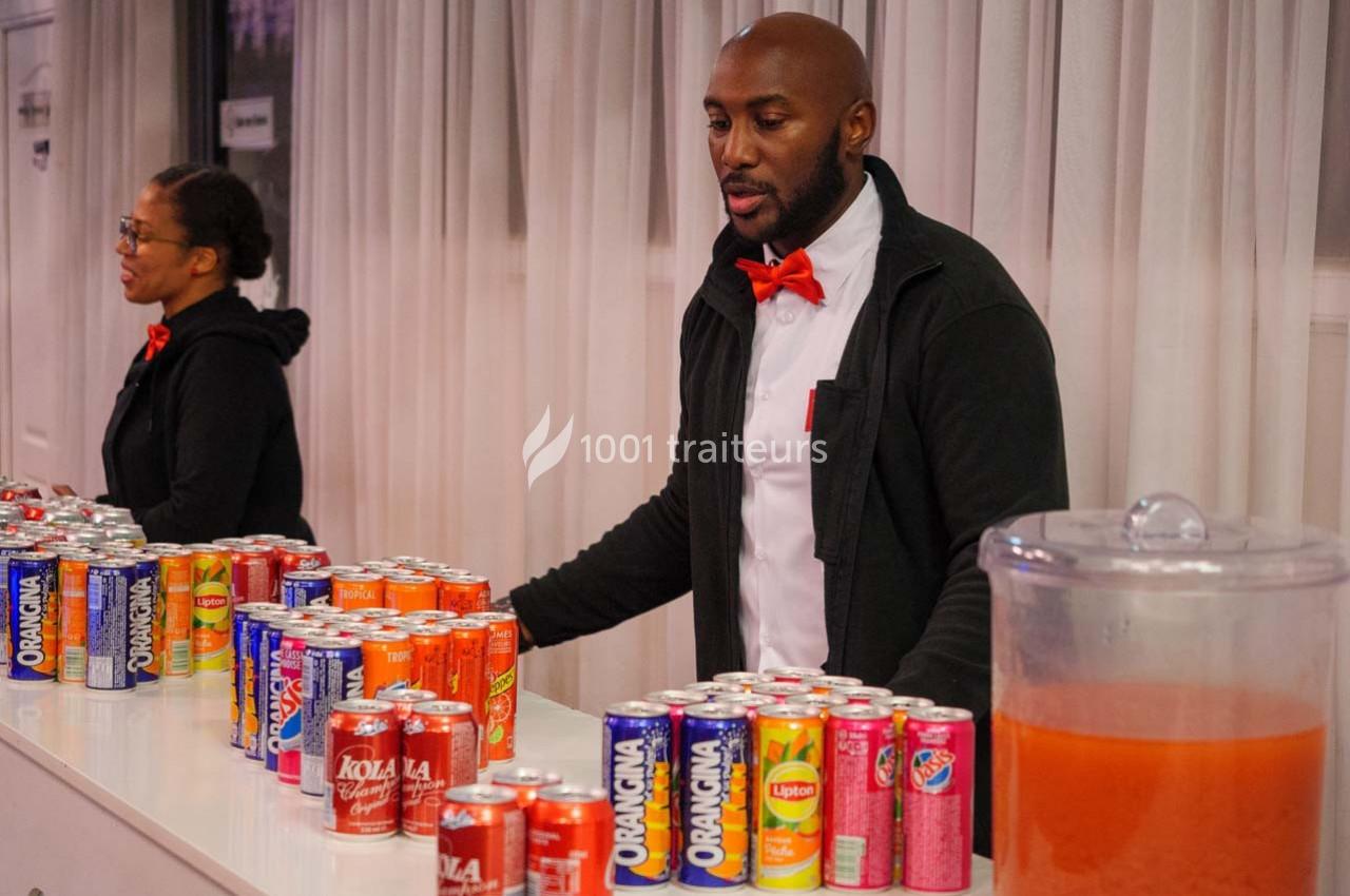 Deux personnes derrière un comptoir proposent des canettes de boissons variées et un récipient de jus.