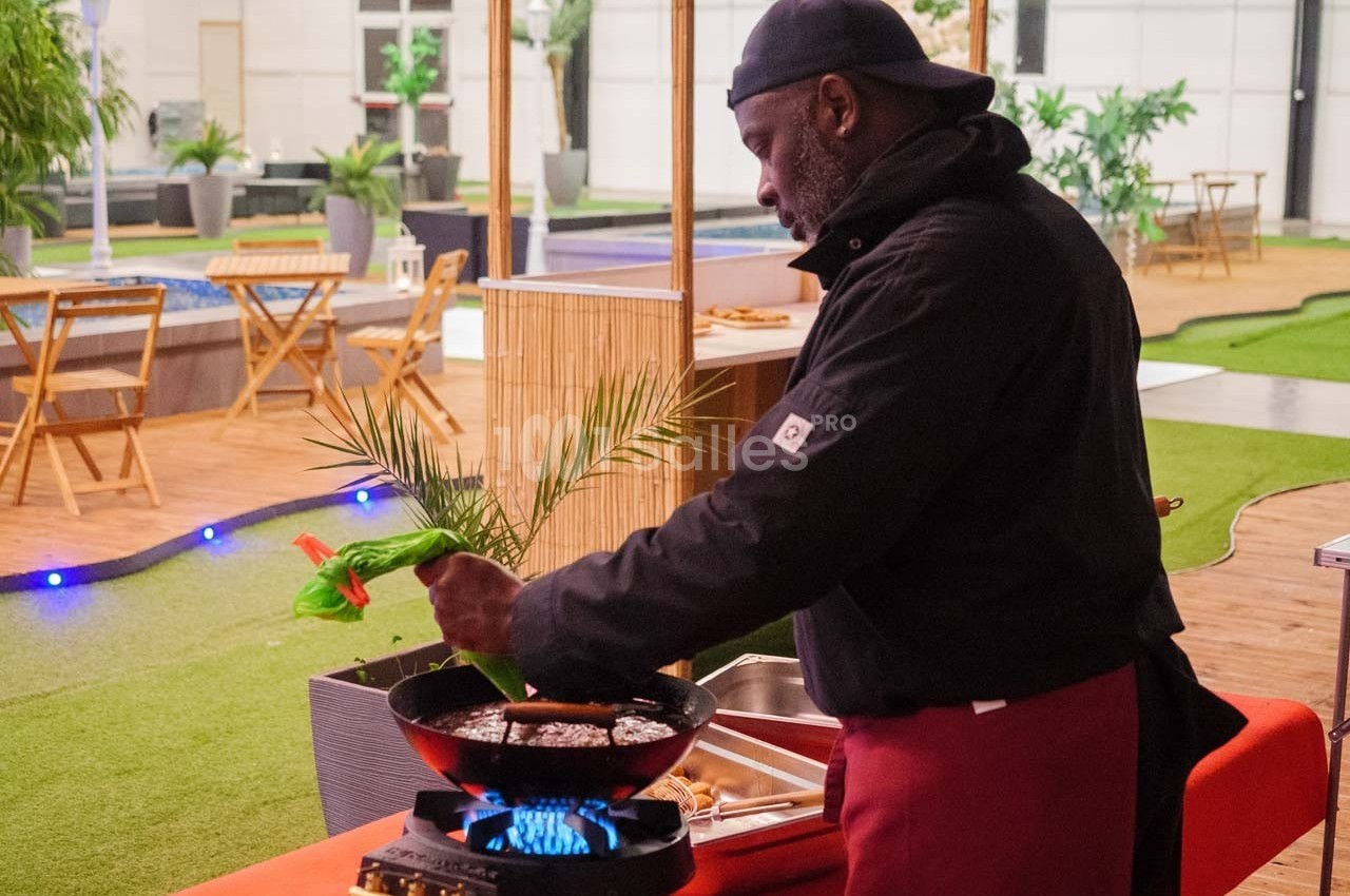Un cuisinier prépare un plat dans une poêle sur un réchaud, dans un espace extérieur aménagé avec des plantes et des tables.