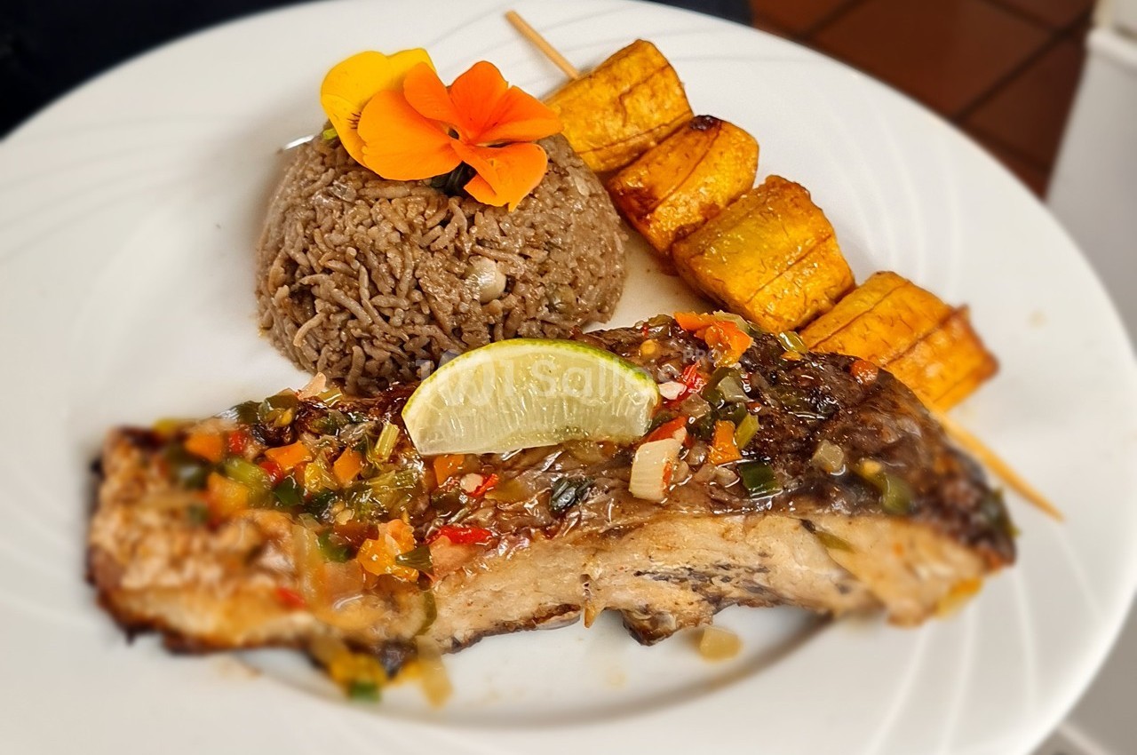 Assiette composée de poisson grillé avec sauce, riz, bananes plantains grillées et décoration florale.