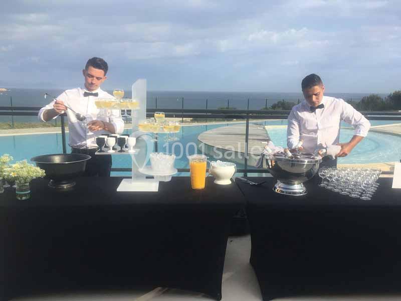 Deux serveurs préparent des boissons sur des tables noires près d'une piscine avec vue sur la mer.
