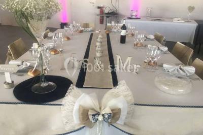 Centre de table floral avec roses et magnolias, bouteille de vin, numéro de table et décorations sur une nappe blanche.