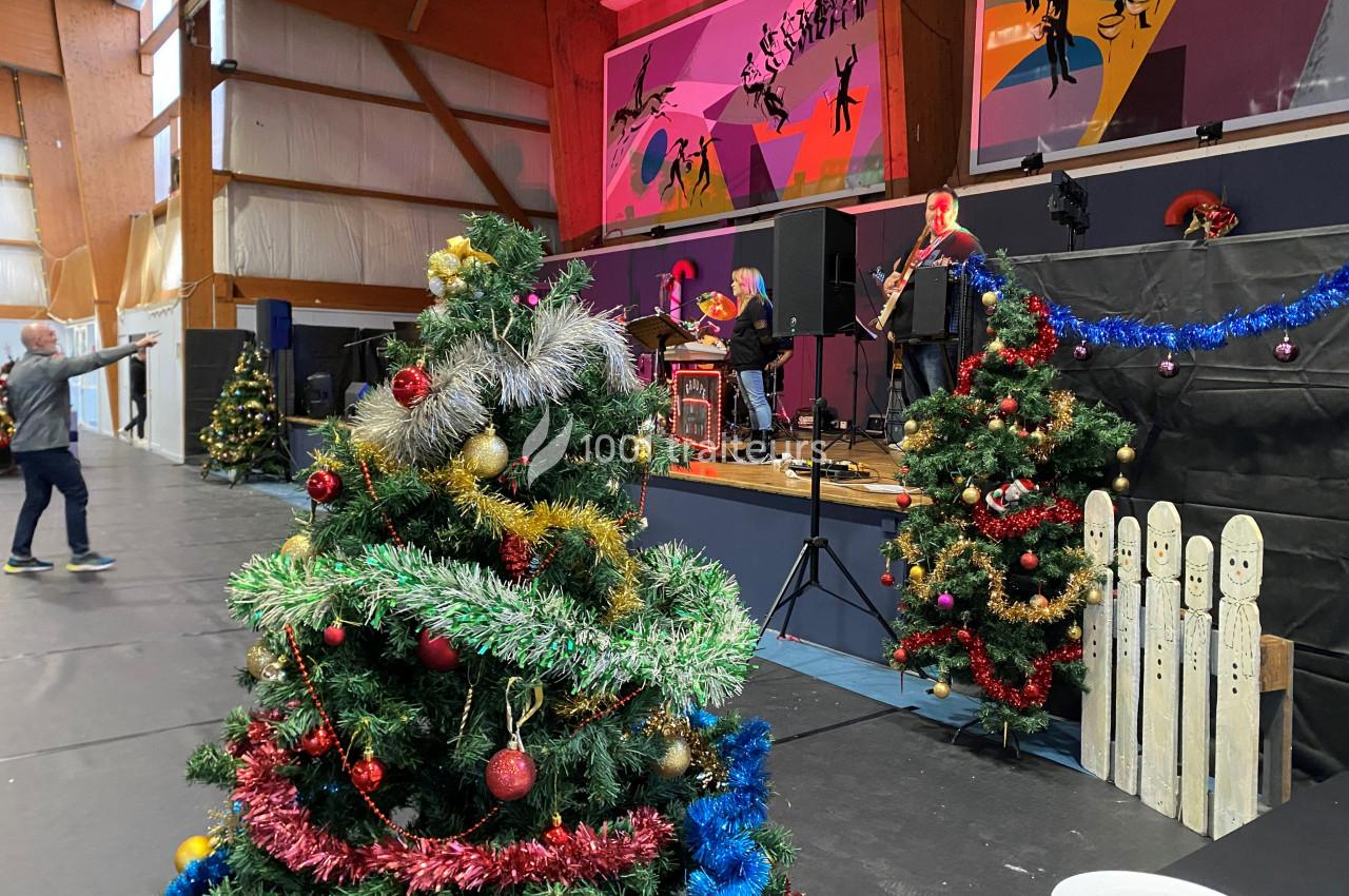 Sapin de Noël décoré au premier plan dans une salle avec scène, musiciens et décorations festives en arrière-plan.