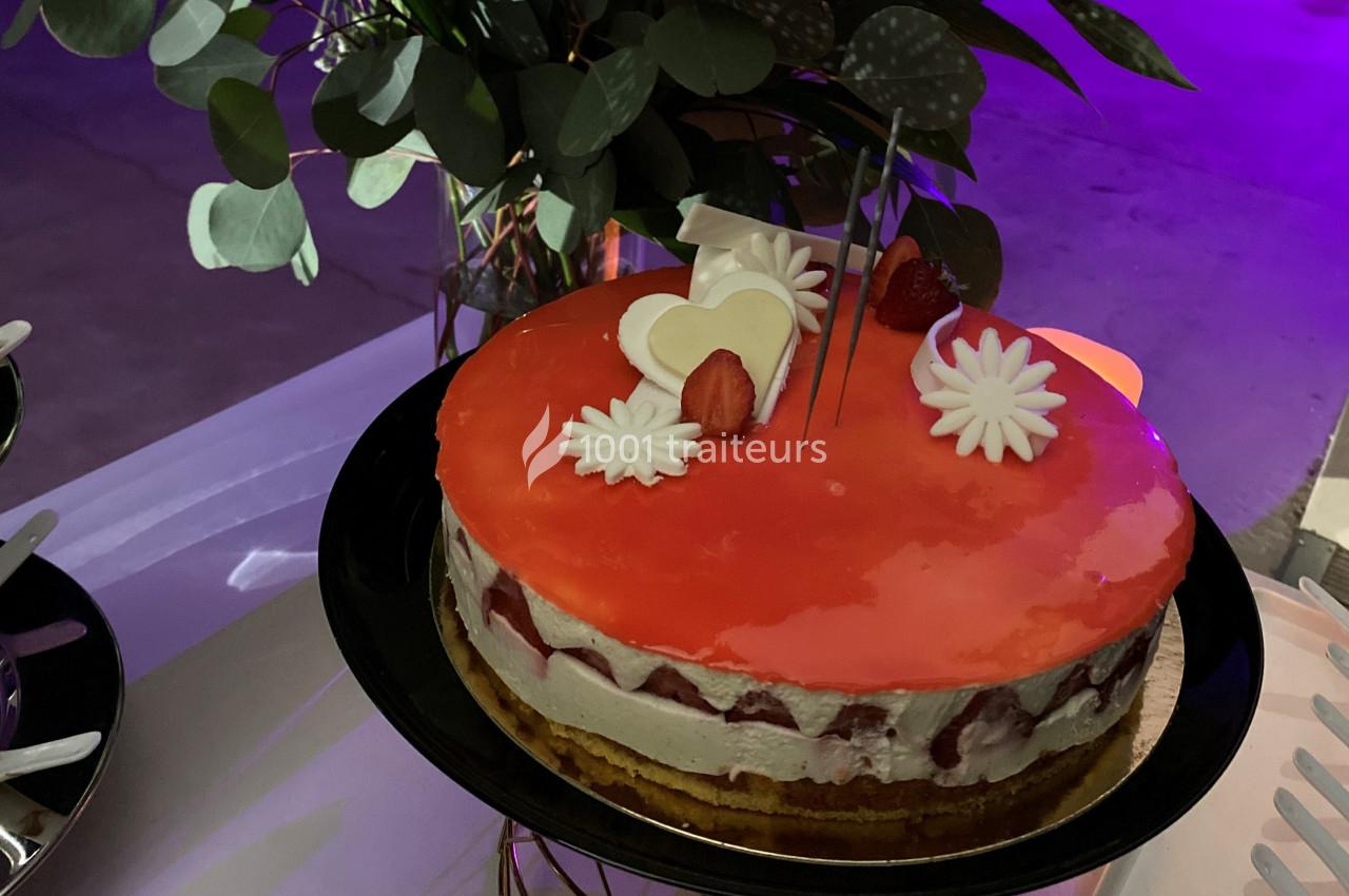 Gâteau avec glaçage rouge, décoré de fraises, fleurs en sucre et un cœur blanc, posé sur une table.