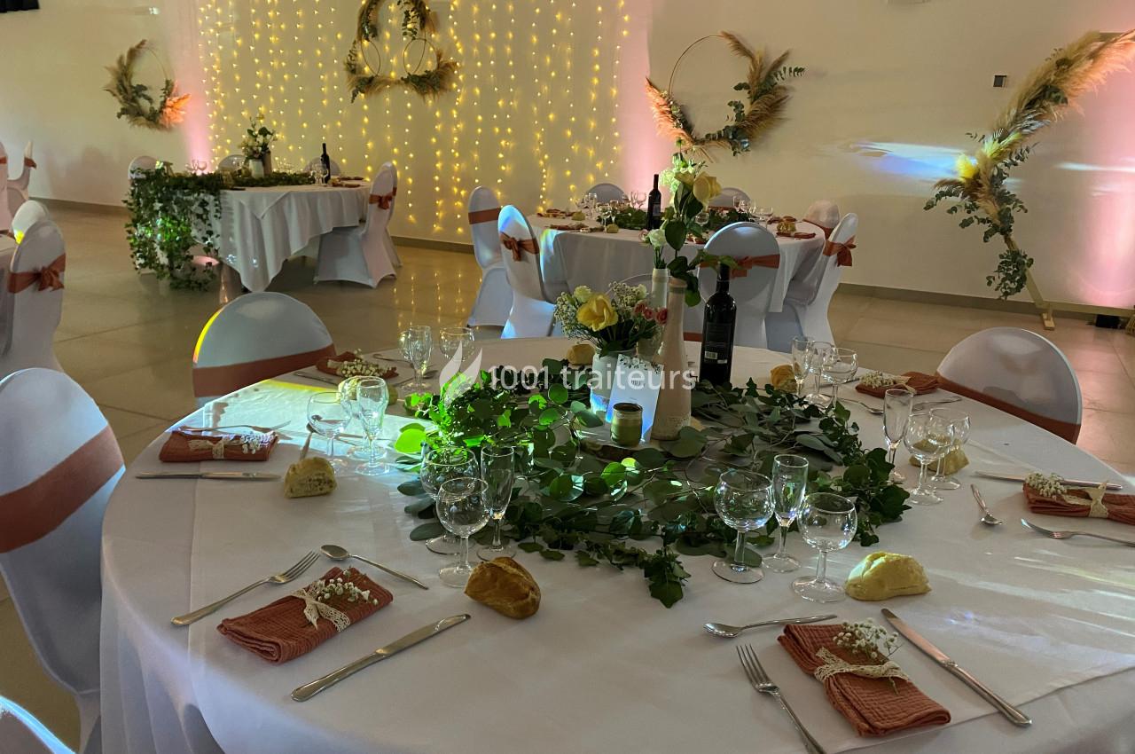 Salle de réception décorée avec des tables rondes dressées, nappes blanches, centres de table végétaux et éclairage…
