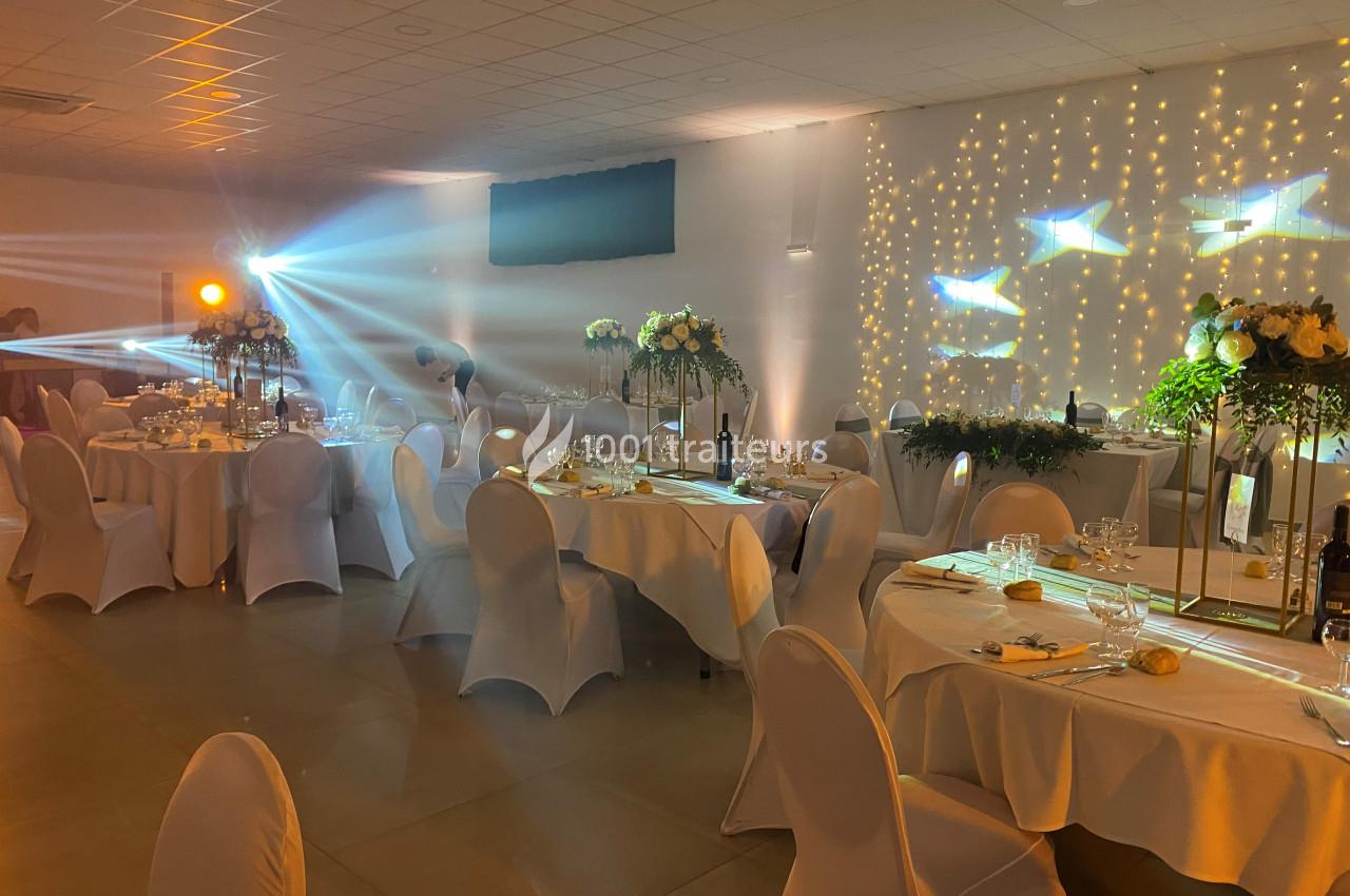 Salle de réception décorée avec des tables rondes, nappes blanches, éclairage tamisé et guirlandes lumineuses au mur.