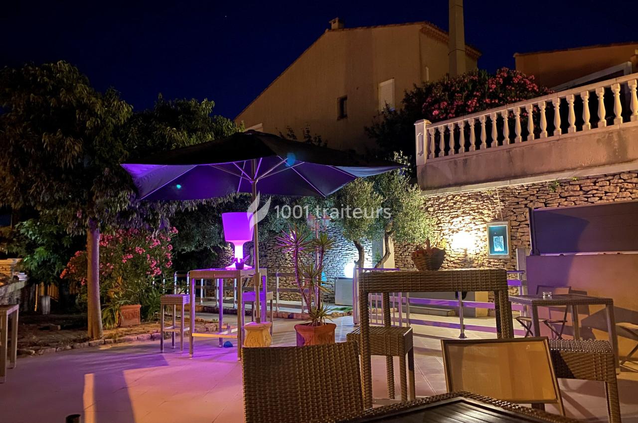 Terrasse extérieure éclairée de nuit avec des tables, des chaises, un grand parasol et des plantes autour.