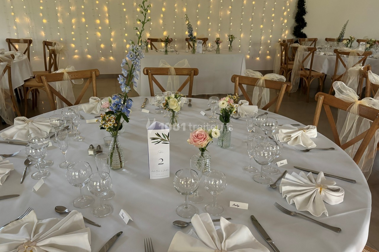 Salle de réception décorée avec des tables rondes, nappes blanches, fleurs et guirlandes lumineuses en arrière-plan.