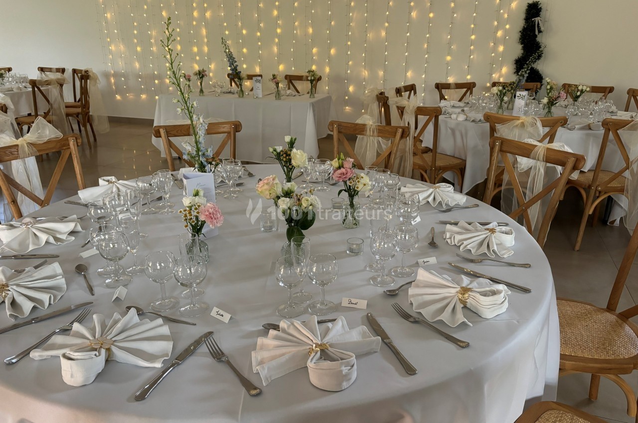 Salle de réception décorée avec des tables rondes dressées, nappes blanches, fleurs et guirlandes lumineuses en arrière-plan.