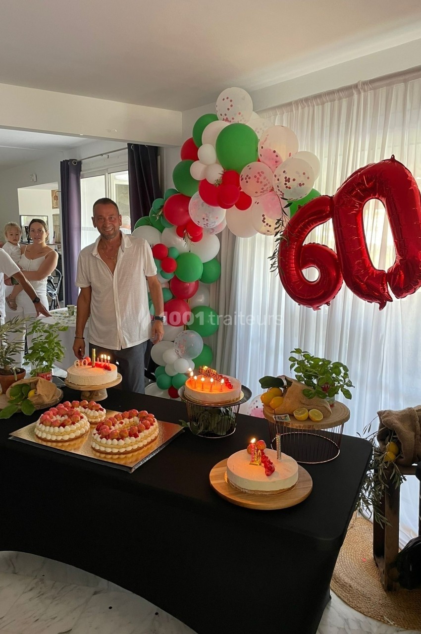 Un homme souriant devant une table décorée avec des gâteaux, des ballons et le chiffre 60 en rouge.