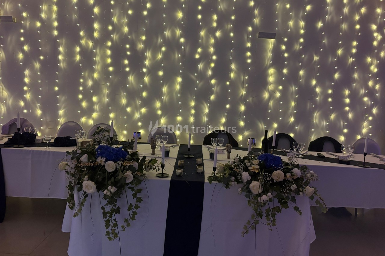 Table décorée pour un événement, ornée de fleurs blanches et bleues, avec un fond de guirlandes lumineuses.