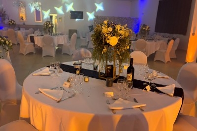 Centre de table floral avec roses et magnolias, bouteille de vin, numéro de table et décorations sur une nappe blanche.