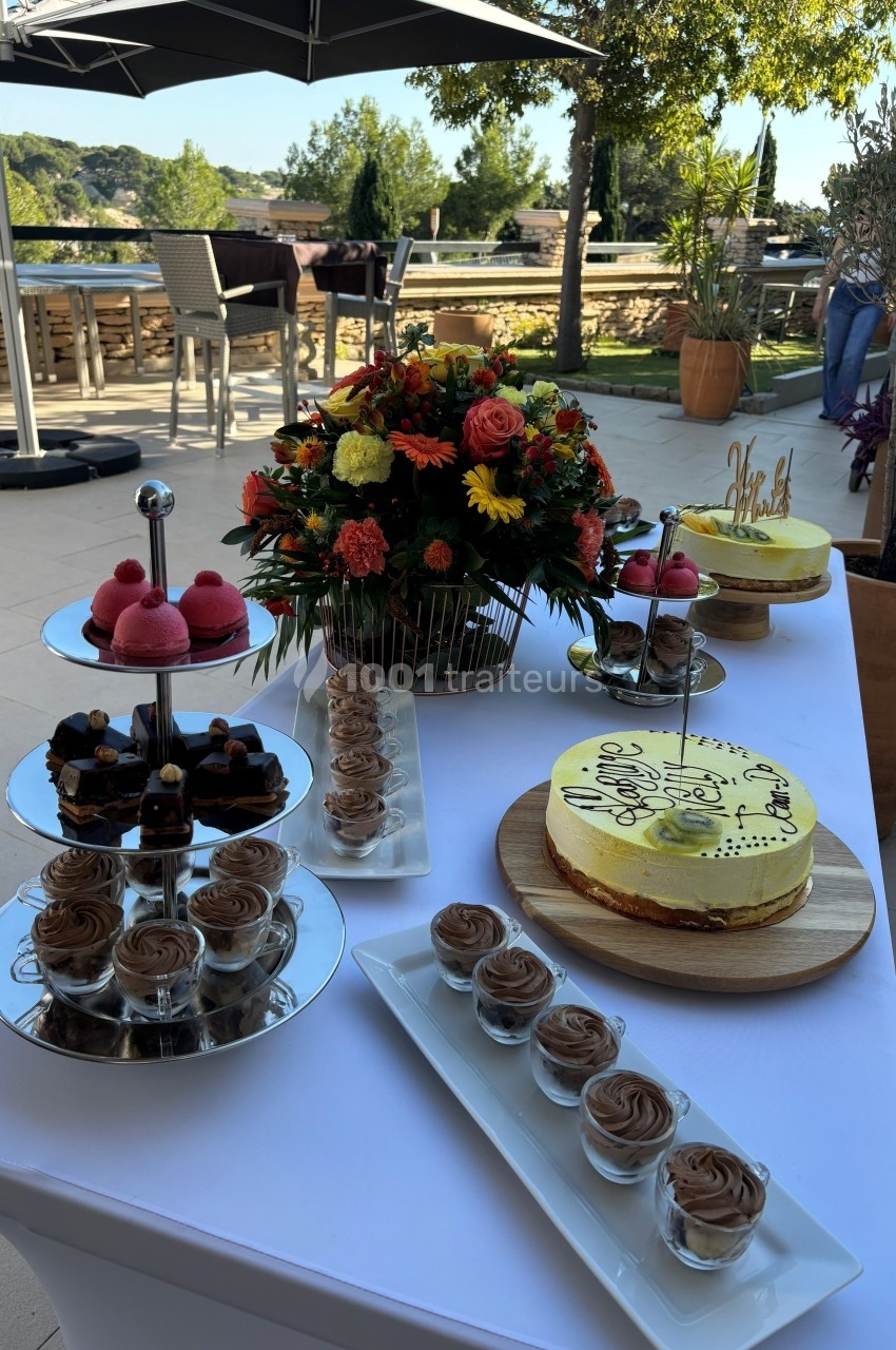 Table dressée en extérieur avec desserts variés, gâteau décoré, fleurs colorées et vue sur terrasse arborée.