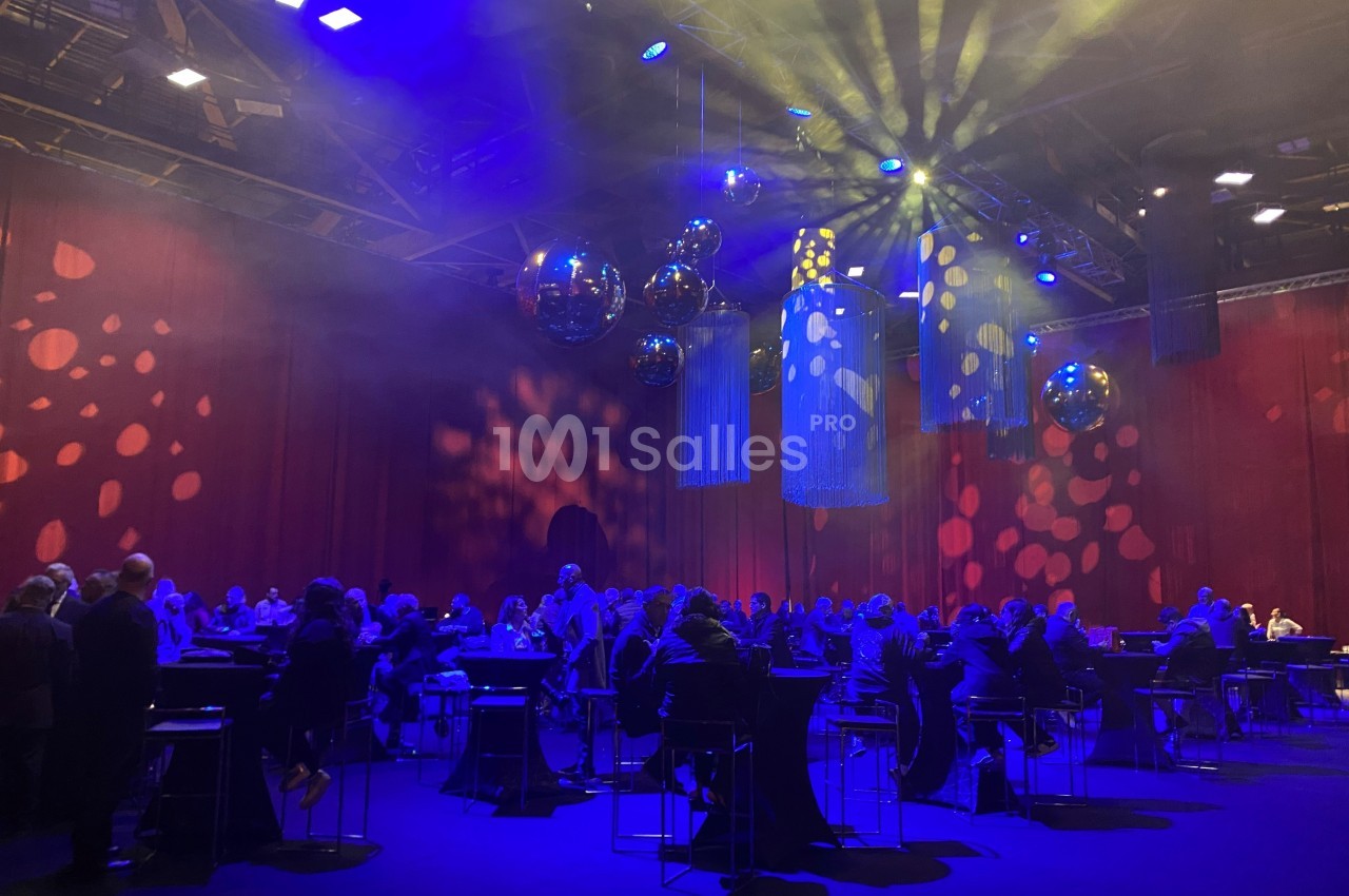 Salle de réception avec invités assis à des tables hautes, éclairée par des lumières colorées et des boules réfléchissantes.