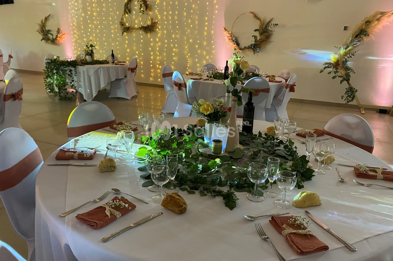 Salle de réception décorée avec des tables rondes dressées, nappes blanches, centres de table végétaux et éclairage…
