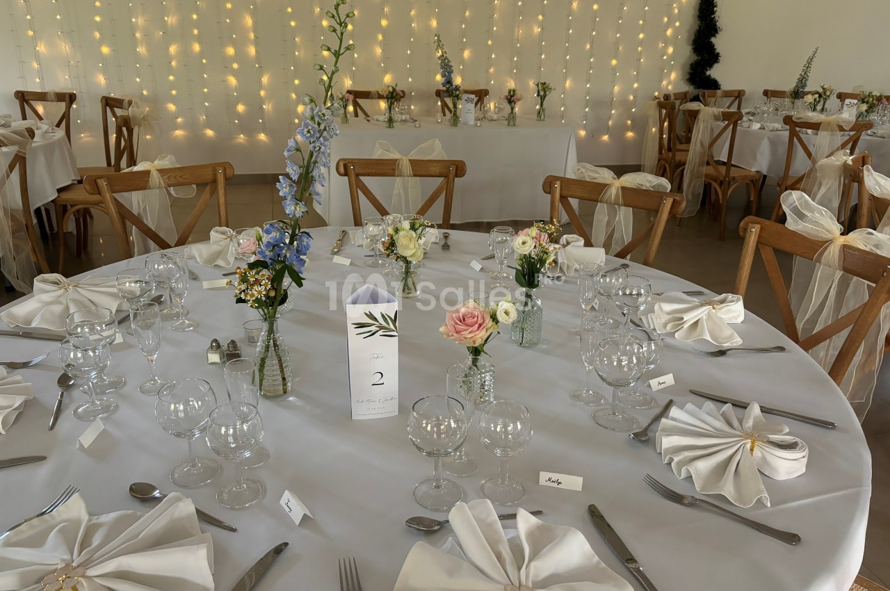 Salle de réception décorée avec des tables rondes, nappes blanches, fleurs et guirlandes lumineuses en arrière-plan.