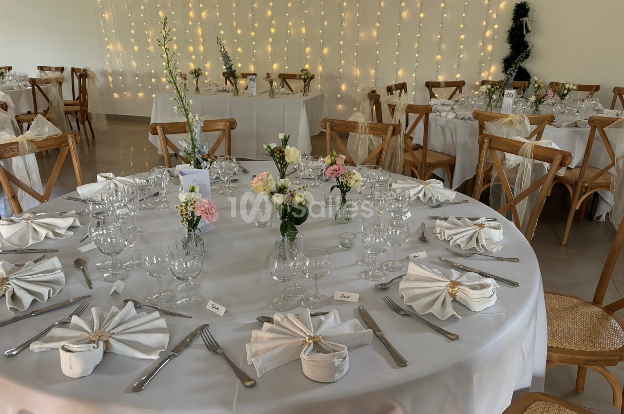 Salle de réception décorée avec des tables rondes dressées, nappes blanches, fleurs et guirlandes lumineuses en arrière-plan.