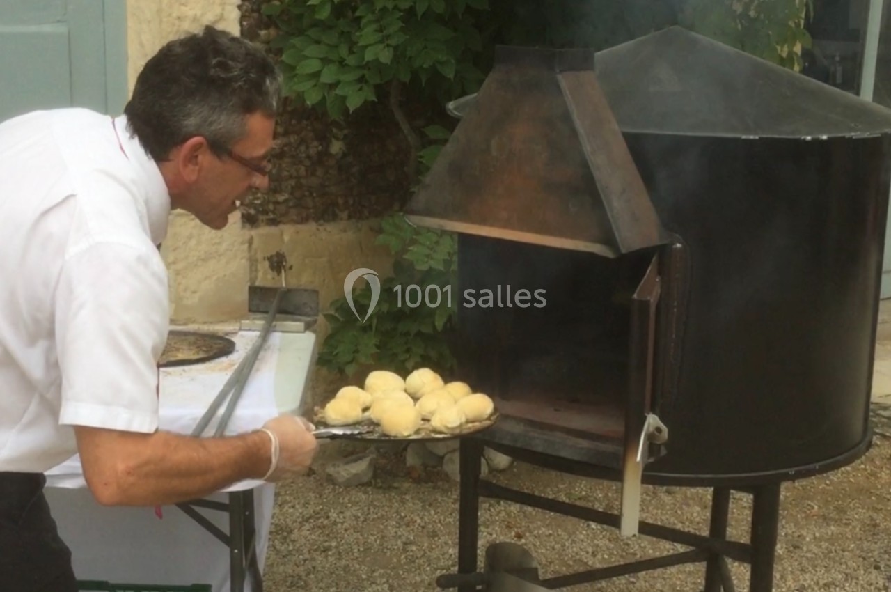 Un homme enfourne des pains ronds dans un four à bois en extérieur, près d'un mur en pierre et de végétation.
